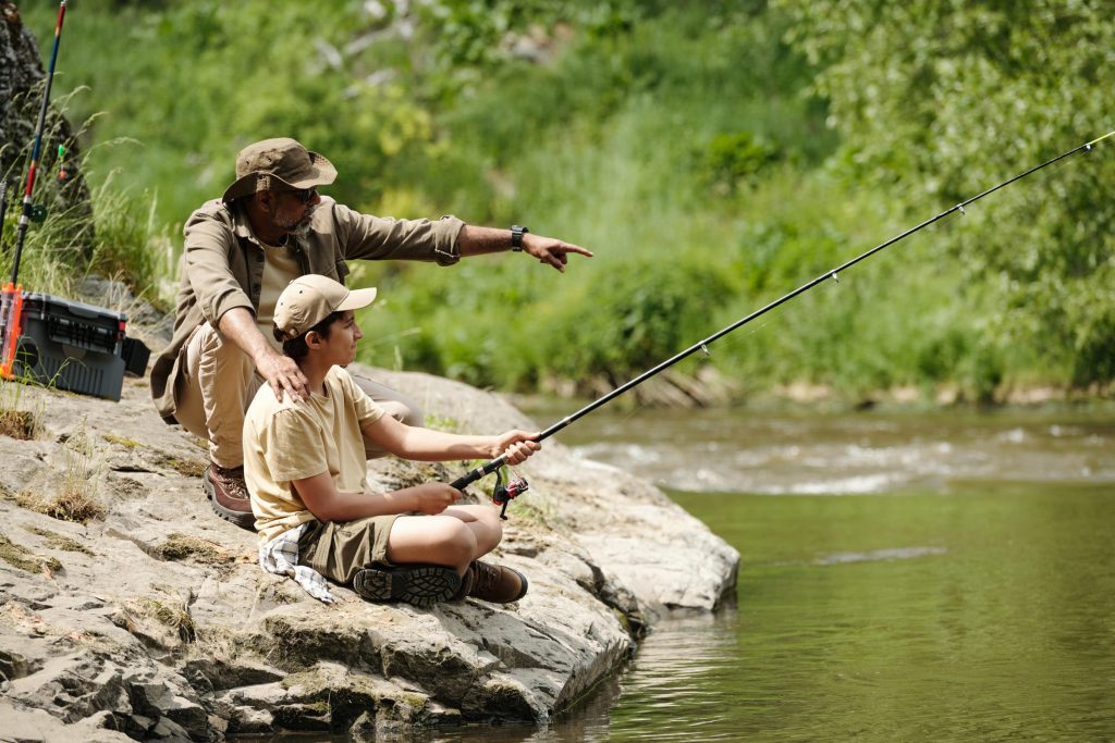 Middle Aged Man Guiding Preteen Boy Fishing by River Outdoors